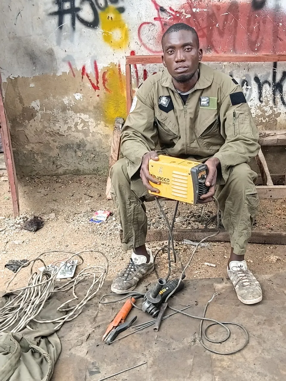 Beka seated in his workshop holding a yellow welding machine